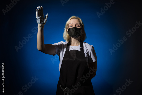 A gardener woman in a black apron, gloves in a protective mask asks and raised her hand up on a dark background.