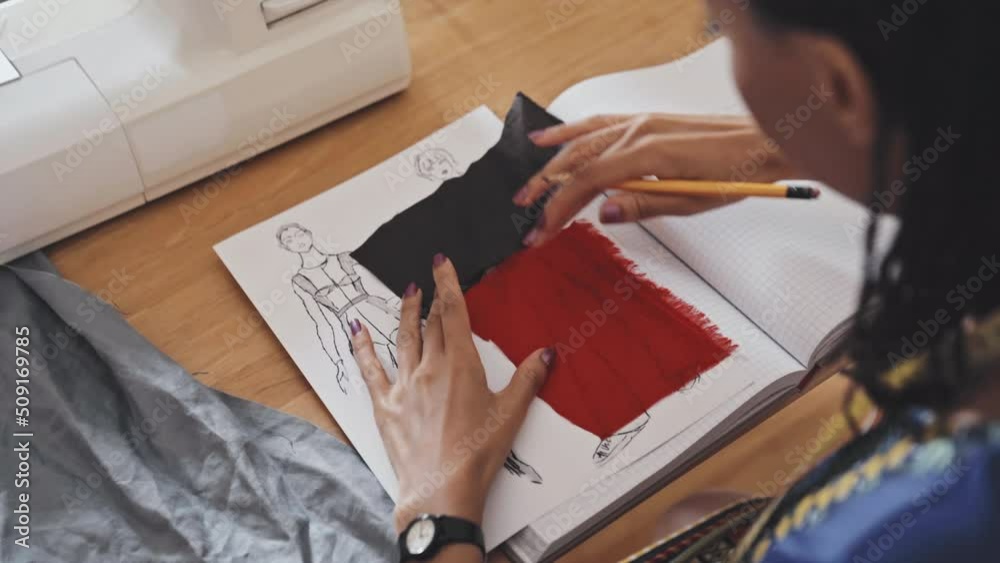 Close up shot of female designer placing two pieces of fabric on fashion sketch while working on new garment collection