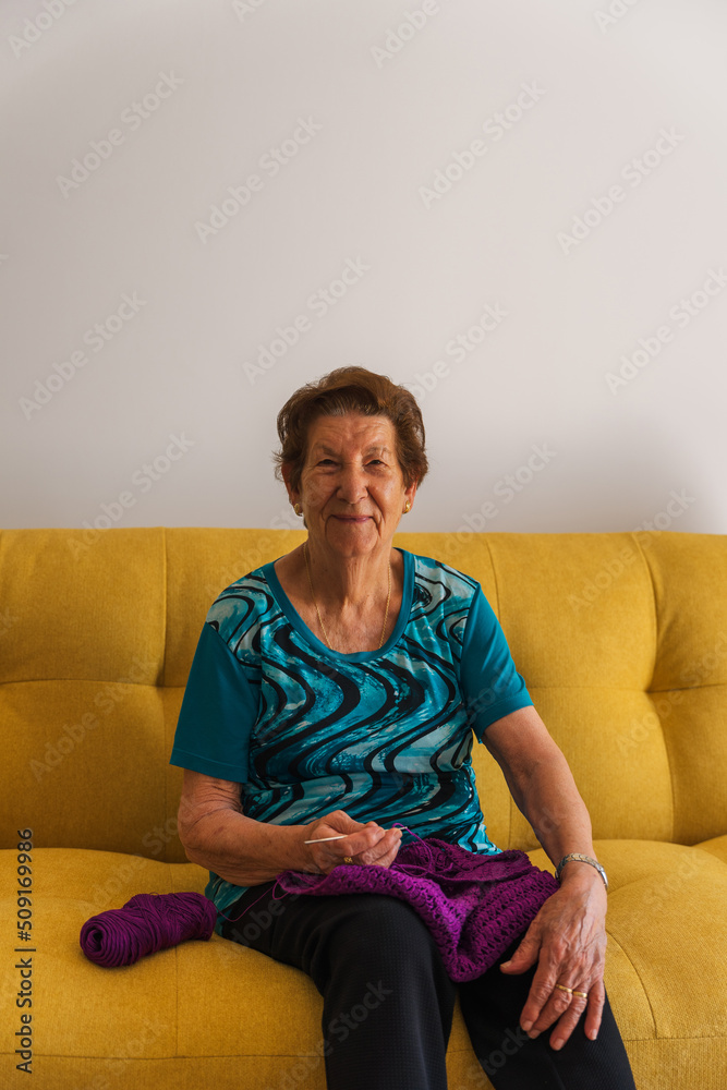 smiling older caucasian woman knitting, looking at the camera. Sitting on a yellow sofa in a blue t-shirt. Grandmother, retired, older,