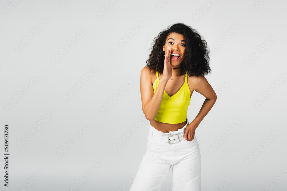 Φωτογραφία happy african american woman in yellow crop top posing with hand in pocket and gossiping isolated on grey