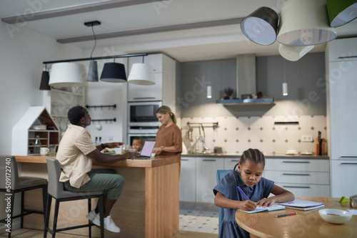 Wide angle portrait of cute black girl studying at kitchen table with multiethnic parents in background, copy space