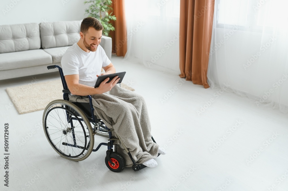 Man in wheelchair using tablet computer at home. Positive retired male with physical disability browsing web on touch pad, watching video online indoors. Man in wheelchair using tablet computer at home. Positive retired male with physical disability browsing web on touch pad, watching video online indoors.