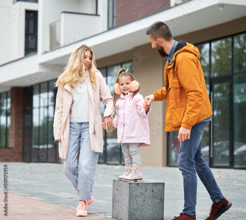 Full length of bearded man and blonde woman playing with little girl on the street in new urban district. Happy loving family with kid having fun outdoors.