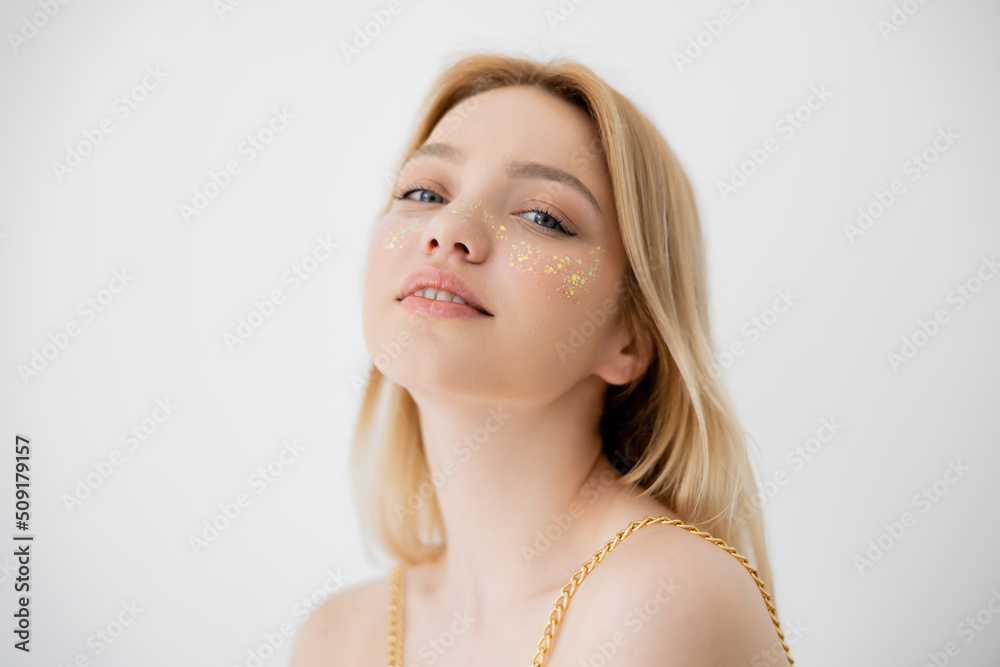 Young woman with glitter on cheeks looking at camera isolated on grey.