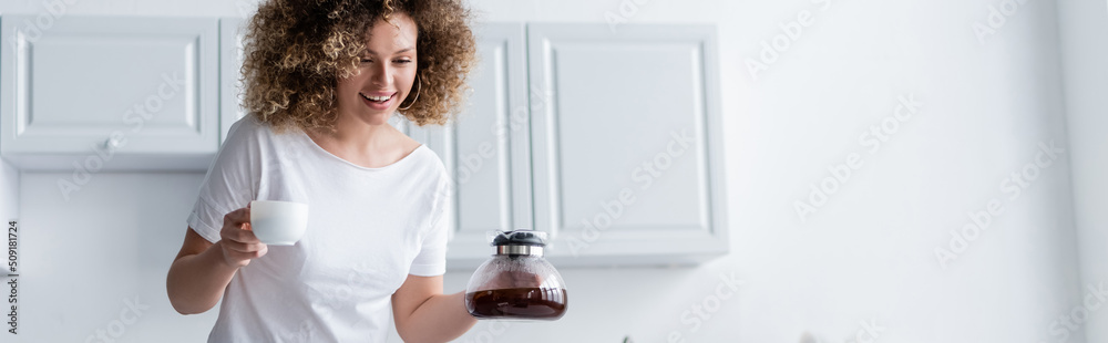 happy woman with wavy hair holding cup and coffee pot in kitchen, banner. happy woman with wavy hair holding cup and coffee pot in kitchen, banner.