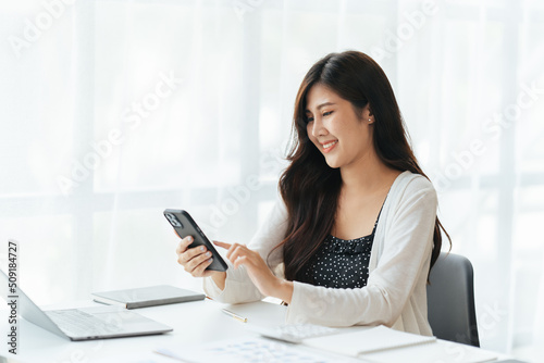 Cheerful young beautiful woman talking on mobile phone and using laptop with smile while sitting at her working place.