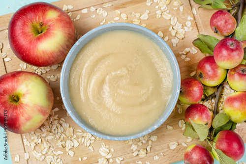 Oatmeal porridge for the baby from ground cereals in a blue bowl, red ripe apples on a wooden board close-up. The first complementary food of a child, baby nutrition.