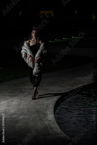 fashion model walking by the side of the pool , black dress and elegant fur, high heels