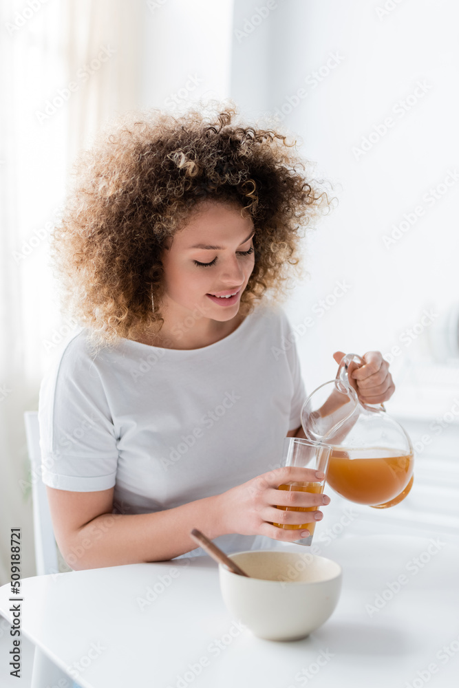 young and positive woman with wavy hair pouring orange juice from jug into glass. young and positive woman with wavy hair pouring orange juice from jug into glass.