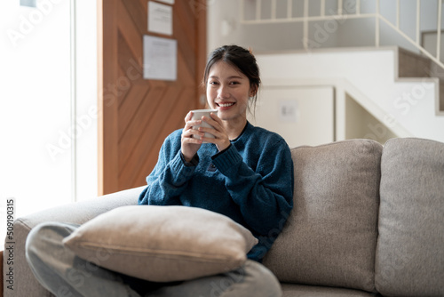 Happy young asian woman sitting on a couch with a cup of coffee at home