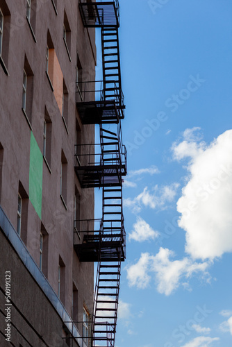 Silhouette of a fire escape on a high-rise building against a blue sky with clouds. Some of the stairs are broken. There is free space for text