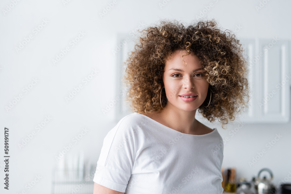 curly woman in white t-shirt smiling at camera in kitchen. curly woman in white t-shirt smiling at camera in kitchen.