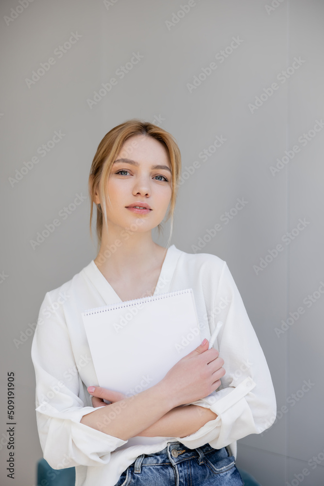 Portrait of young woman in blouse holding notebook and looking at camera at home. Portrait of young woman in blouse holding notebook and looking at camera at home.