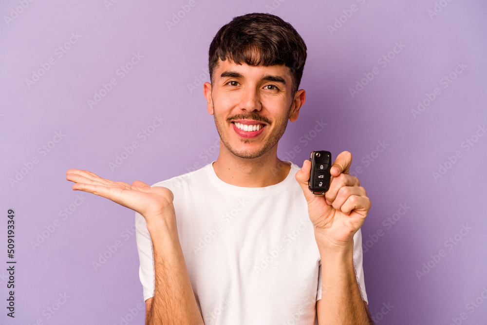 Young hispanic man holding keys car isolated on purple background showing a copy space on a palm and holding another hand on waist. Young hispanic man holding keys car isolated on purple background showing a copy space on a palm and holding another hand on waist.