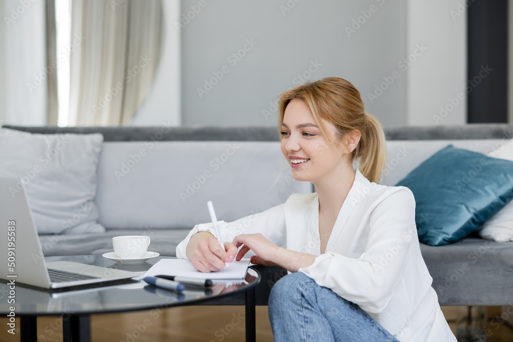 Positive young freelancer writing on notebook near laptop on coffee table at home.