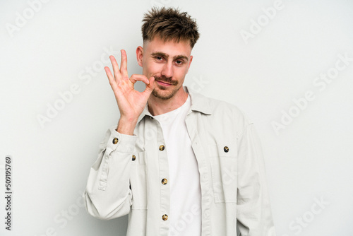 Young caucasian man isolated on white background winks an eye and holds an okay gesture with hand.
