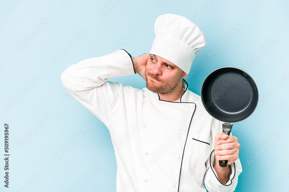 Young caucasian chef man holding flying pan isolated on blue background touching back of head, thinking and making a choice. Young caucasian chef man holding flying pan isolated on blue background touching back of head, thinking and making a choice.