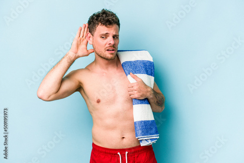 Young caucasian man holding beach towel isolated on blue background trying to listening a gossip.