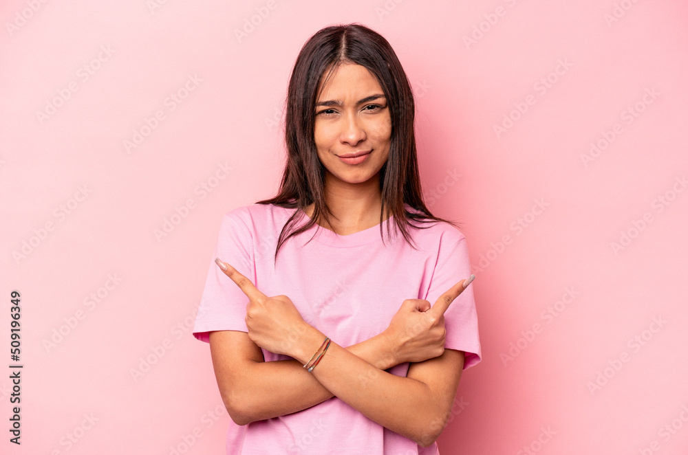 Young hispanic woman isolated on pink background points sideways, is trying to choose between two options.