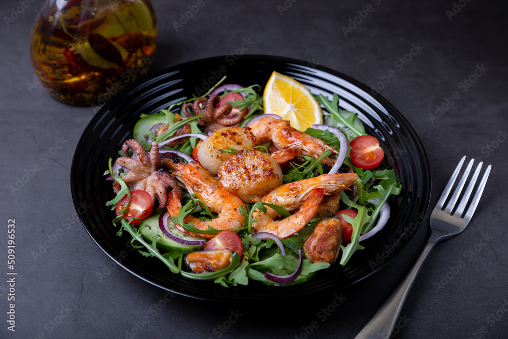 Salad with seafood, arugula, tomatoes, cucumbers, red onion and lemon on a black plate. Black background, close-up. Salad with seafood, arugula, tomatoes, cucumbers, red onion and lemon on a black plate. Black background, close-up.