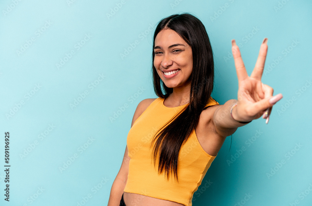 Fototapeta premium Young hispanic woman isolated on blue background joyful and carefree showing a peace symbol with fingers.