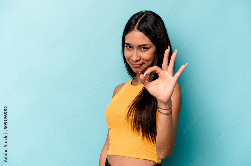 Fototapeta premium Young hispanic woman isolated on blue background winks an eye and holds an okay gesture with hand.