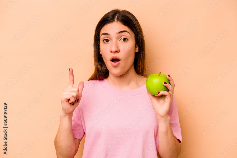 Young hispanic woman holding an apple isolated on beige background having an idea, inspiration concept. Young hispanic woman holding an apple isolated on beige background having an idea, inspiration concept.