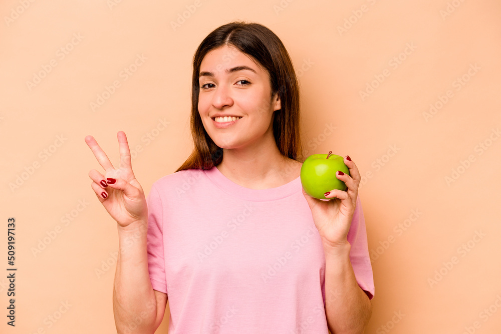 Young hispanic woman holding an apple isolated on beige background showing number two with fingers. Young hispanic woman holding an apple isolated on beige background showing number two with fingers.