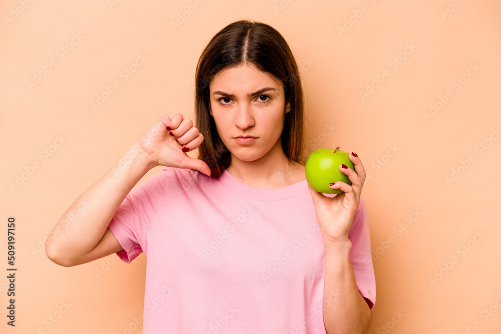 Young hispanic woman holding an apple isolated on beige background showing a dislike gesture, thumbs down. Disagreement concept. Young hispanic woman holding an apple isolated on beige background showing a dislike gesture, thumbs down. Disagreement concept.