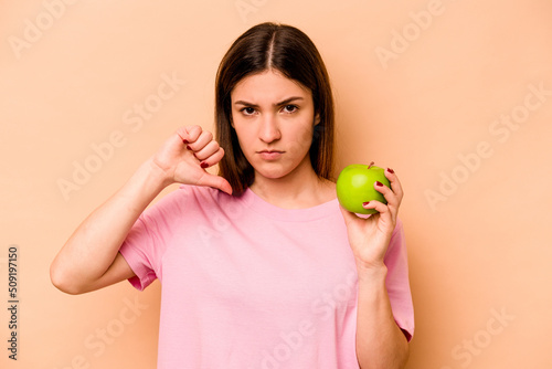 Young hispanic woman holding an apple isolated on beige background showing a dislike gesture, thumbs down. Disagreement concept. © Asier