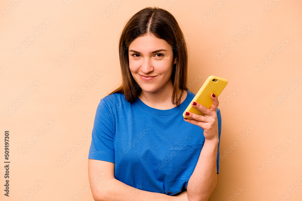 Young caucasian woman holding mobile phone isolated on beige background laughing and having fun. Young caucasian woman holding mobile phone isolated on beige background laughing and having fun.