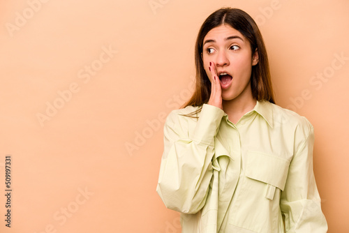 Young hispanic woman isolated on beige background is saying a secret hot braking news and looking aside