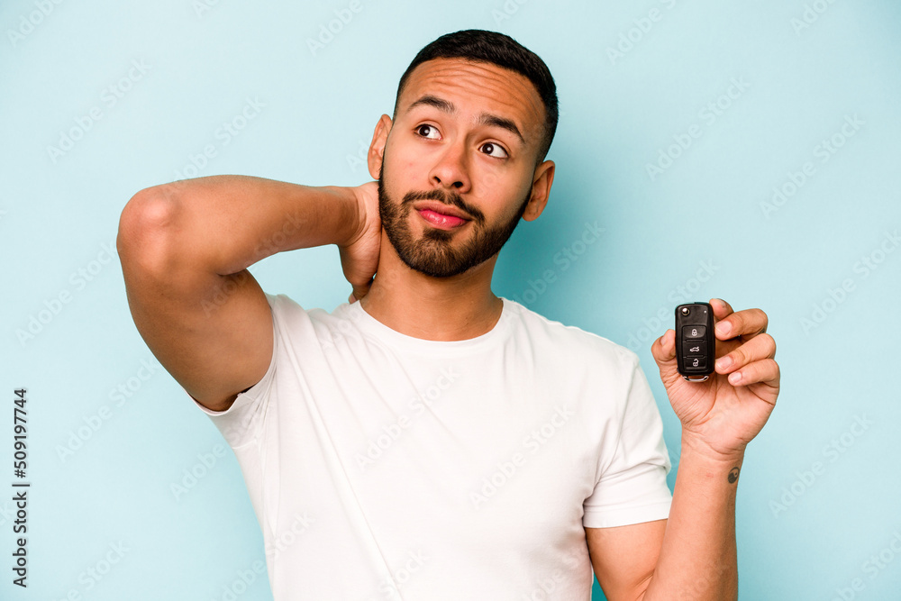 Young hispanic man holding car keys isolated on blue background touching back of head, thinking and making a choice. Young hispanic man holding car keys isolated on blue background touching back of head, thinking and making a choice.