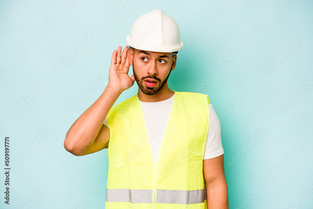 Young laborer hispanic man isolated on blue background trying to listening a gossip.