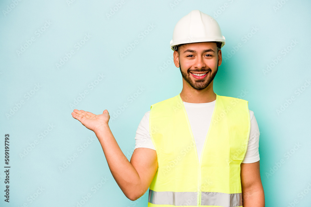 Young laborer hispanic man isolated on blue background showing a copy space on a palm and holding another hand on waist. Young laborer hispanic man isolated on blue background showing a copy space on a palm and holding another hand on waist.