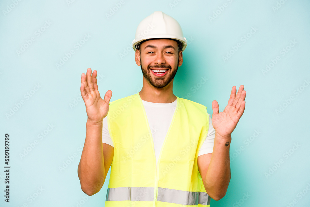 Young laborer hispanic man isolated on blue background receiving a pleasant surprise, excited and raising hands. Young laborer hispanic man isolated on blue background receiving a pleasant surprise, excited and raising hands.