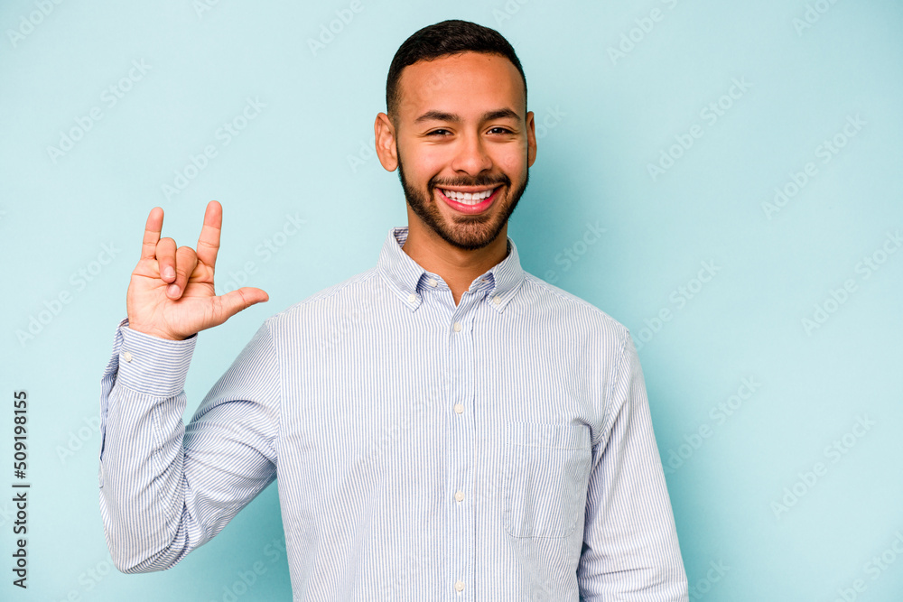 Young hispanic man isolated on blue background showing a horns gesture as a revolution concept. Young hispanic man isolated on blue background showing a horns gesture as a revolution concept.