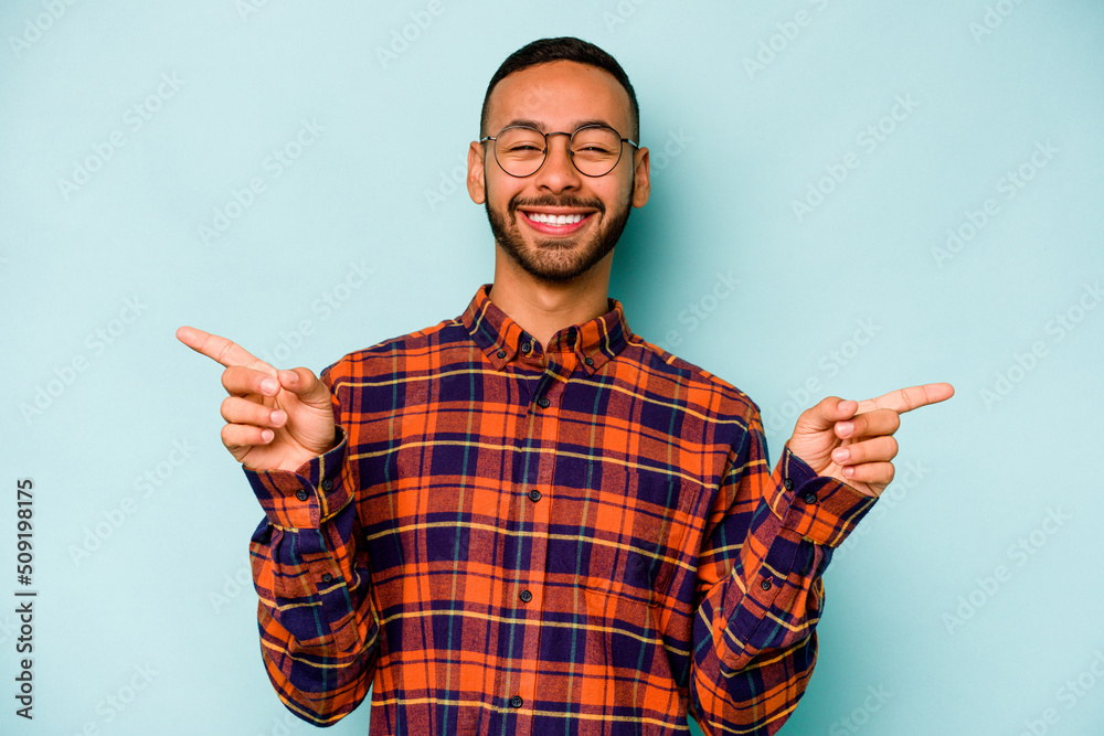 Young hispanic man isolated on blue background pointing to different copy spaces, choosing one of them, showing with finger. Young hispanic man isolated on blue background pointing to different copy spaces, choosing one of them, showing with finger.