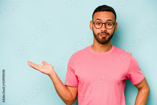 Young hispanic man isolated on blue background showing a copy space on a palm and holding another hand on waist.