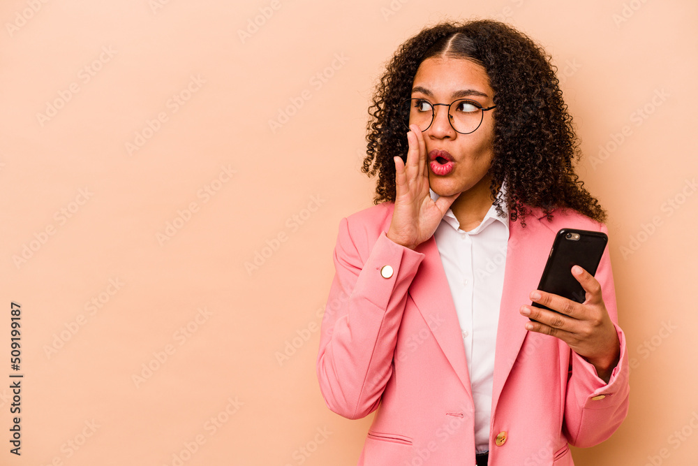 Young African American woman holding mobile phone isolated on beige background is saying a secret hot braking news and looking aside Young African American woman holding mobile phone isolated on beige background is saying a secret hot braking news and looking aside