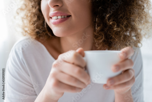 partial view of smiling woman with curly hair holding white coffee cup.