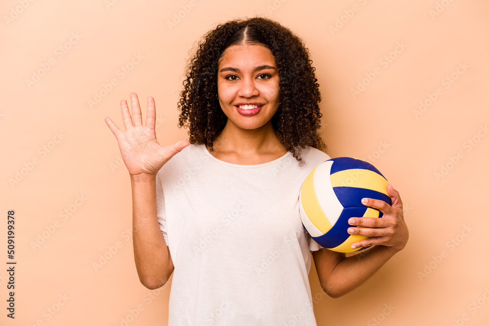Young African American woman playing volleyball isolated on beige background smiling cheerful showing number five with fingers.