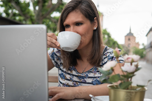 Brunette woman sipping a delicious freshly brewed coffee while using her laptop. Young woman with a smile doing business or work online with her computer. Concept of telecommuting. photo