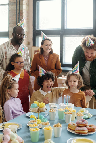 Little boy in party hat blowing candles on birthday cake while sitting at table with dessert and celebrating birthday with adults and children