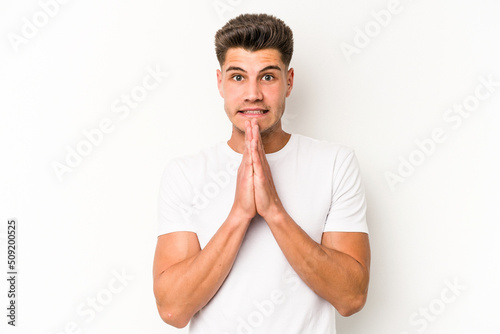Young caucasian man isolated on white background holding hands in pray near mouth, feels confident.