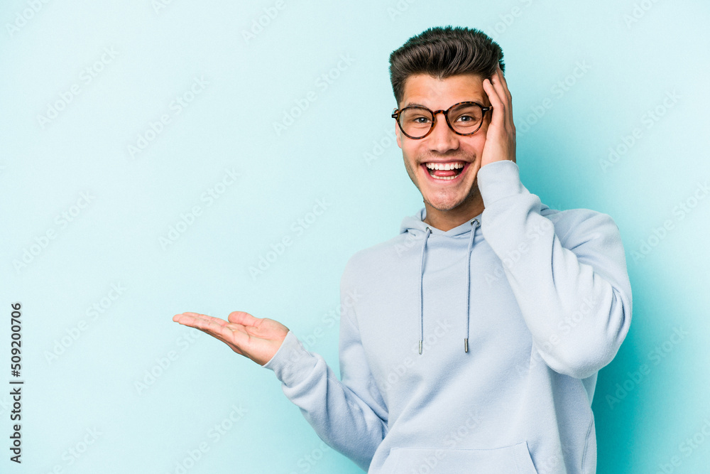 Young caucasian man isolated on blue background holds copy space on a palm, keep hand over cheek. Amazed and delighted. Young caucasian man isolated on blue background holds copy space on a palm, keep hand over cheek. Amazed and delighted.