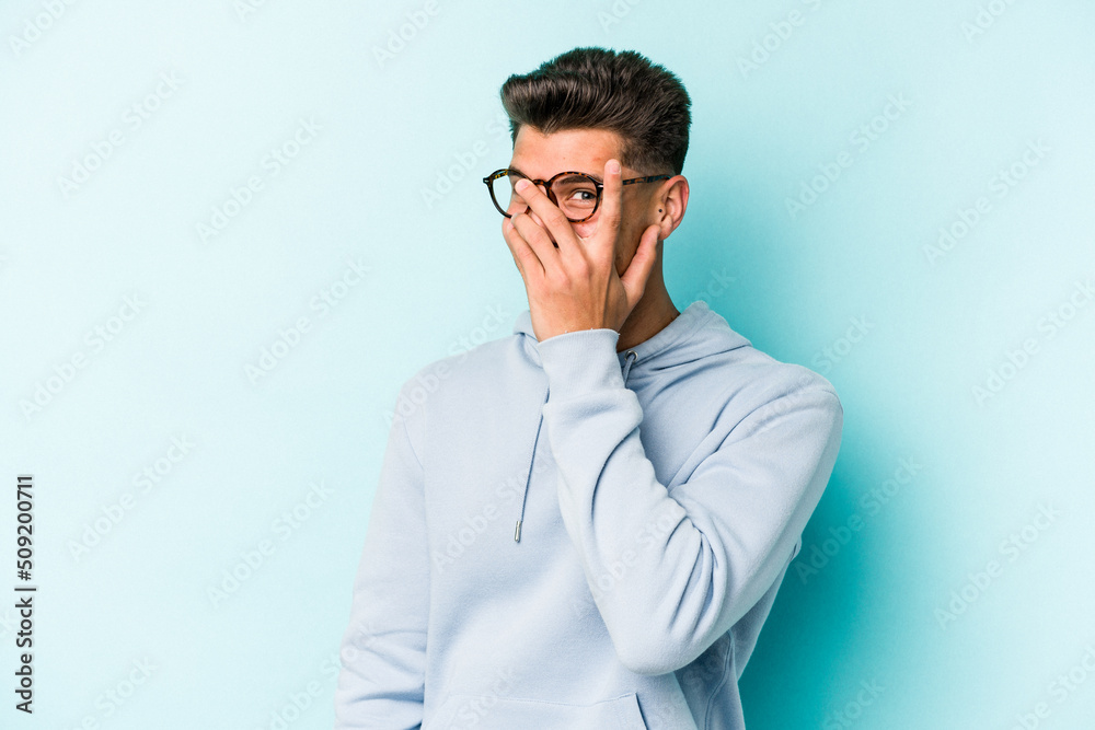 Young caucasian man isolated on blue background blink at the camera through fingers, embarrassed covering face. Young caucasian man isolated on blue background blink at the camera through fingers, embarrassed covering face.