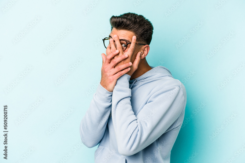 Young caucasian man isolated on blue background blink through fingers frightened and nervous. Young caucasian man isolated on blue background blink through fingers frightened and nervous.