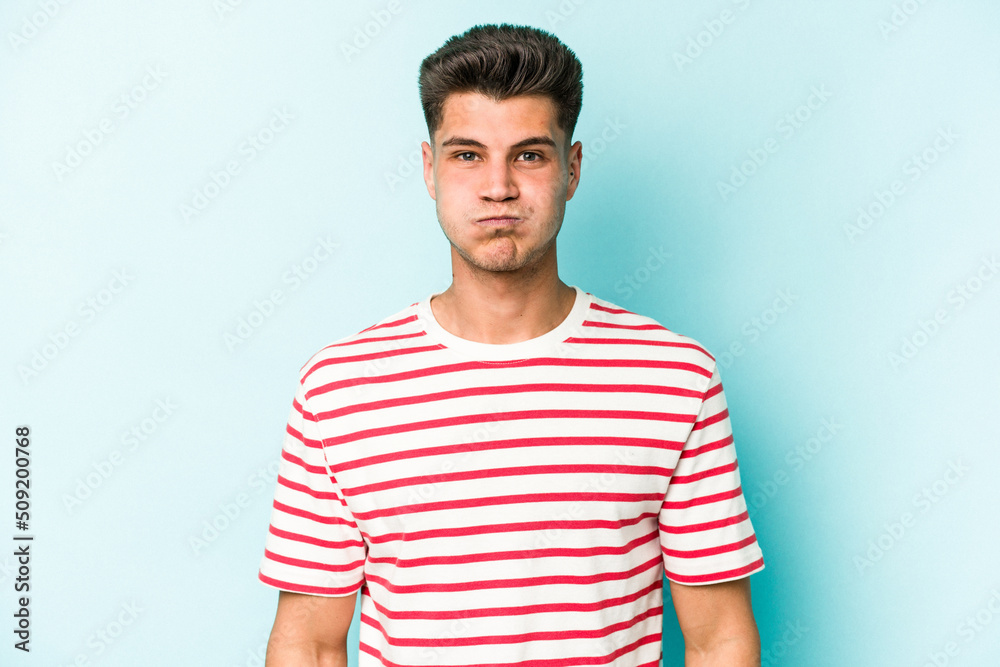 Young caucasian man isolated on blue background blows cheeks, has tired expression. Facial expression concept.