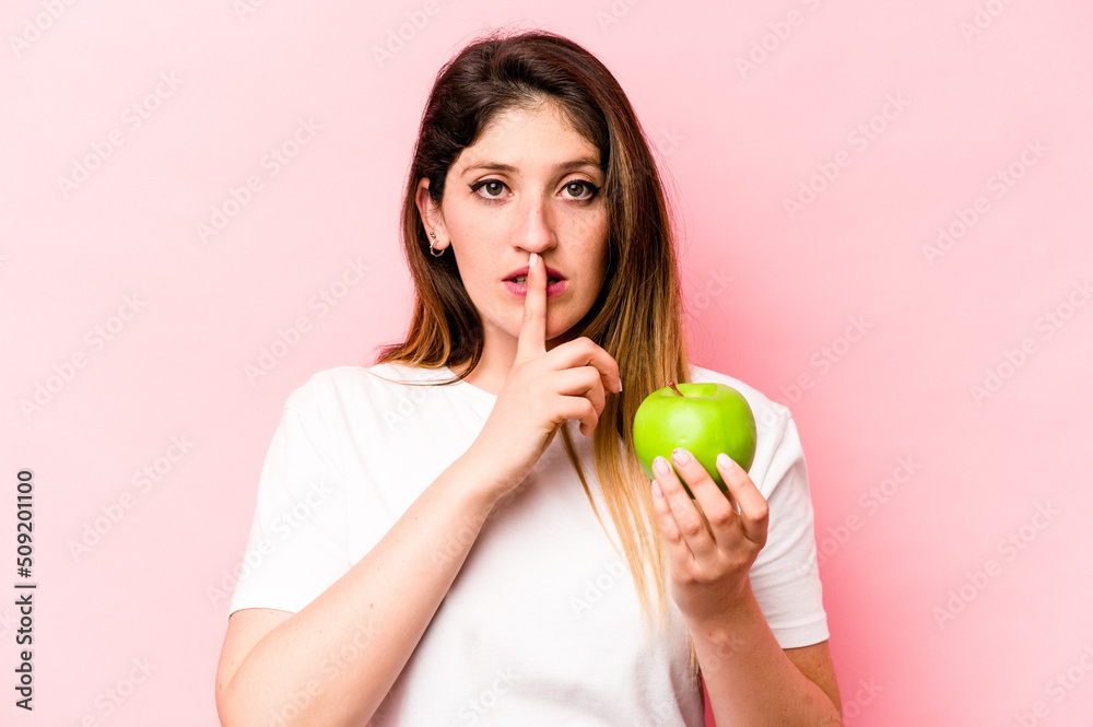 Young caucasian woman holding an apple isolated on pink background keeping a secret or asking for silence. Young caucasian woman holding an apple isolated on pink background keeping a secret or asking for silence.
